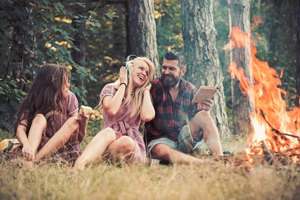 Friends relax at bonfire flame with sparks in vintage style. People camping at fire in forest. Women and bearded man at campfire. Eating food, reading book and entertainment. Summer vacation concept