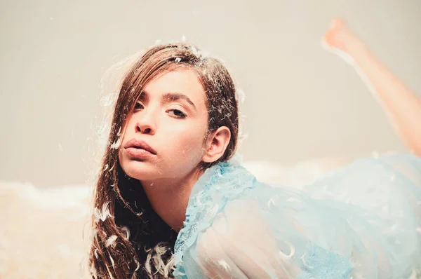 Woman with long hair in tender pajama. Lady in transparent blue nightie lay on bed while feathers fly around. Tenderness concept. Girl on calm face lay on bed covered with feathers and fluff. — Stock Photo, Image