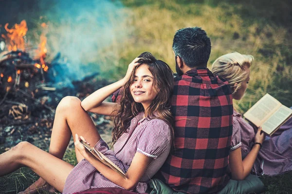 Camping in wilderness. Turn back guy looking at fire while two beautiful girls read book. Smiling brunette with braces leaning on her friend