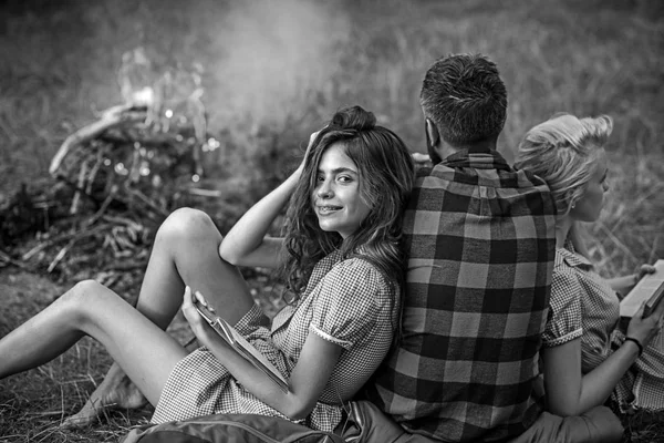 Vrienden kamperen in het bos. Weer man kijkt bij kampvuur, terwijl twee meisjes op hem leunen. Brunette meisje glimlachend met accolades lezen boek — Stockfoto