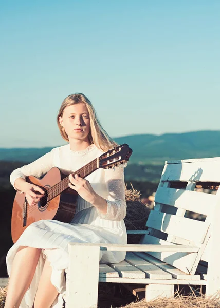 Chica albina sostener la guitarra acústica, instrumento de cuerda. Músico de moda en vestido blanco en la naturaleza soleada. Mujer sensual tocar la guitarra en el banco de madera. Mujer guitarrista realizar concierto de música — Foto de Stock