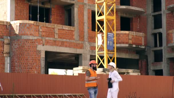 Två unga ingenjörer går genom byggarbetsplats, bakgrund stor kran, de bär säkerhetsutrustning. Arbetar på byggarbetsplatsen. Konstruktion koncept. — Stockvideo