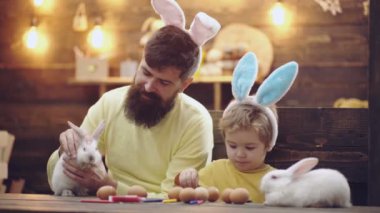 Mutlu aile Paskalya için hazırlıyoruz. Sevimli küçük çocuk çocuk bunny kulaklar giyiyor. Yumurta boyama güzel çocuk çocuk. Baba ve oğul Paskalya yumurtaları boya. Paskalya tavşanı ahşap bir masa üzerinde.