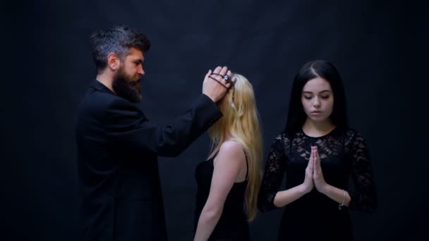 Priester zegent de meisjes. Concept van religie. Bebaarde man in zwart en twee meisjes bidden op zwarte achtergrond. Religie is een opium voor het volk. — Stockvideo