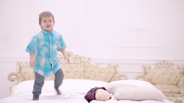 Niño con el pelo rubio saltando en la cama, divirtiéndose en el dormitorio. Niño pequeño jugando en la cama, casa de ocio. Niño es feliz y divertido en el dormitorio familiar . — Vídeos de Stock