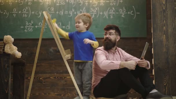 眼鏡の笑顔の先生は子供が黒板を拭くのを見ます。父と興奮したブロンドの子供は数学を学びます。サイドビューの男の子と男は、ラップトップと床に座っています。木製の背景にチョークで描く少年. — ストック動画