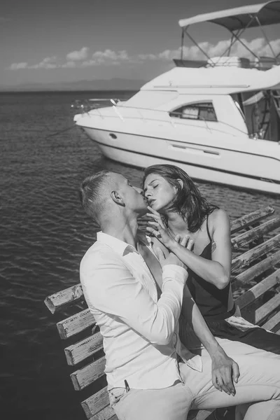 Woman and man in fashionable clothes full of desire near luxury yacht. Honeymoon concept. Couple in love on honeymoon. Young passionate couple sit on bench at dock near boat and kiss, sunny summer day