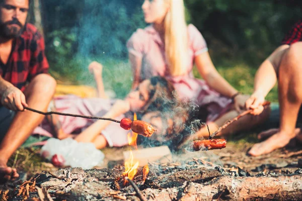 Cozinhar salsichas em paus sobre chamas de fogueira, acampar durante a noite no verão. Grupo de amigos sentados à fogueira — Fotografia de Stock