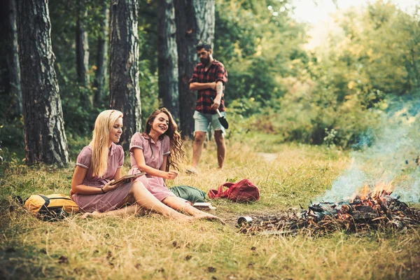 レトロなドレスのファッションの女性は焚き火でおくつろぎください。夏の森でぼやけた男と幸せな女性は。長い髪の美しさの女の子は、緑の草に笑顔します。休暇やキャンプ旅行と読書本、ヴィンテージ — ストック写真