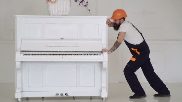 Homem com barba trabalhador no capacete e macacão empurra, esforços para mover piano. Carregador move instrumento de piano. Homem com barba, trabalhador de macacão e capacete adormecer cansado, fundo branco . — Vídeo de Stock
