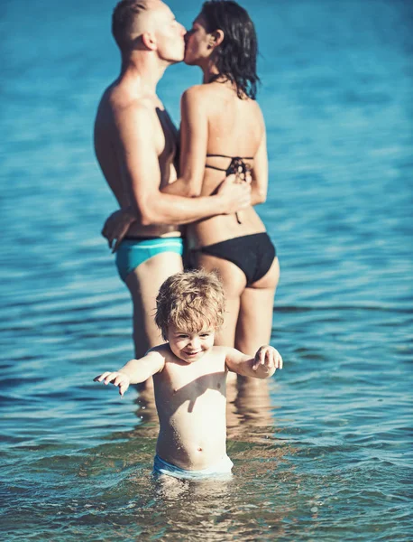 Un niño con un padre y una madre besando. Viajar en familia con el niño el día de la madre o el padre. Vacaciones de verano de familia feliz. El amor y la confianza como valores familiares. Madre y padre con hijo en el mar en la playa . — Foto de Stock