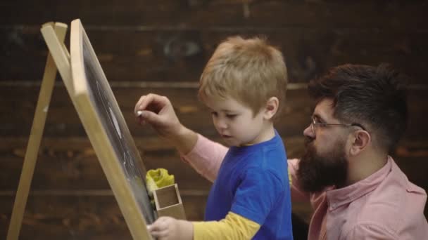 Padre e figlio dipinti con gesso su tavola. Vista laterale di bambino e padre con pezzi di gesso disegno quadro su lavagna isolato su sfondo di legno. Felice famiglia dipinge gesso . — Video Stock