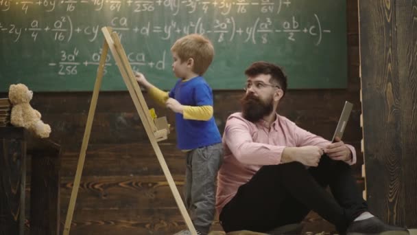 Le professeur vérifie l'écriture des élèves sur un tableau. Elève parler à son tuteur ou papa tout en résolvant la tâche. Concept d'enseignement à domicile. Professeur souriant dans des lunettes assis sur le sol pendant que l'enfant s'amuse . — Video