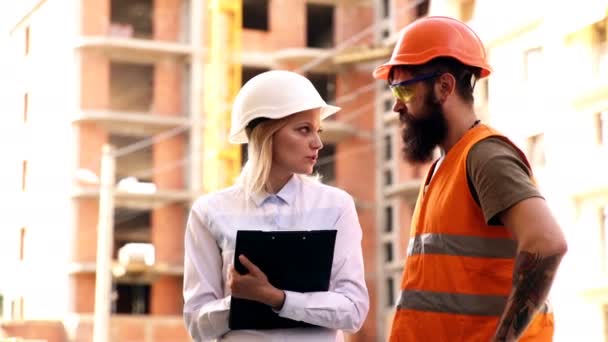 Två civilingenjörer hålla planer och inspektera byggarbetsplatsen. Manliga och kvinnliga builder i Skyddshjälmar kommunicera på bakgrunden av en byggnad under uppförande. — Stockvideo