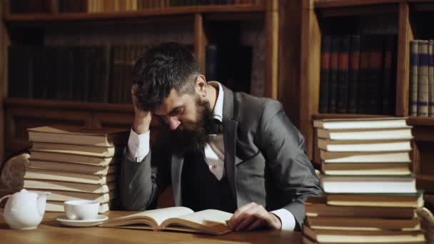 Estudiante Hipster estudiando en la biblioteca. Investigación y estudio del concepto. Hombre en traje clásico, profesor con la cara ocupada concentrada se sienta en la biblioteca cerca de montones de libros, estanterías en el fondo . — Vídeo de stock