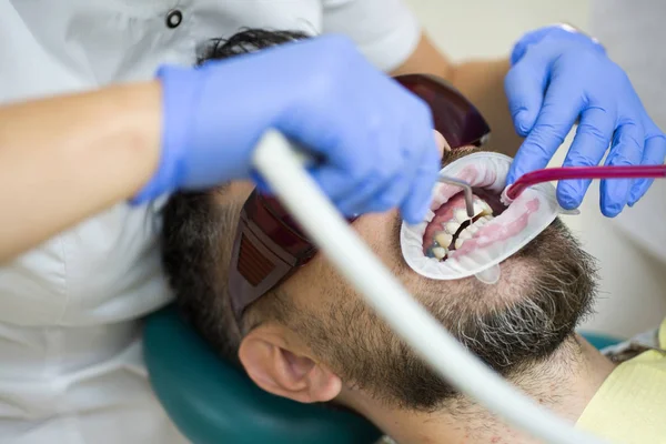 Jeune homme choisissant la couleur des dents chez le dentiste. Dentiste examinant les dents des patients à la clinique. Jeune homme souriant après la visite dentiste sur fond flou lumineux . — Photo