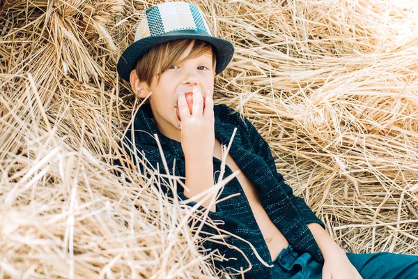 Little boy advertises natural products. Happy child at the autumn fair. Portrait of a cheerful boy lying in a hay. Fair-haired boy lies on hay background and eats an apple.