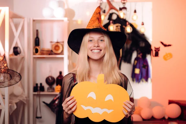 Halloween Witch in black hat. Woman posing with Pumpkin. Indoor portrait of cute young witch at halloween party. Pretty young blonde woman dressed as a fairy with pumpkin. Helloween people concept.