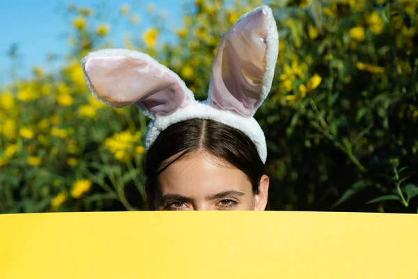 Hermosa joven con orejas de conejo y póster en blanco sobre fondo exterior. Copiar espacio. Concepto de vacaciones de Pascua. Descuento . — Foto de Stock