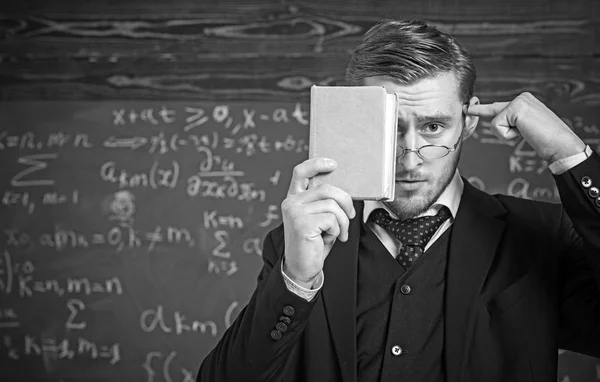 Retrato de primer plano del estudiante sosteniendo el libro frente a su cara y apuntando a su cabeza con otra mano. Pensativo hombre inteligente en traje de pie en el aula —  Fotos de Stock