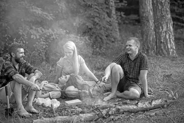 Smiling friends having picnic in woods. Two happy couples enjoying summer evening outdoors, romantic date. Bearded man and his friend frying sausages over fire while their girlfriends rest