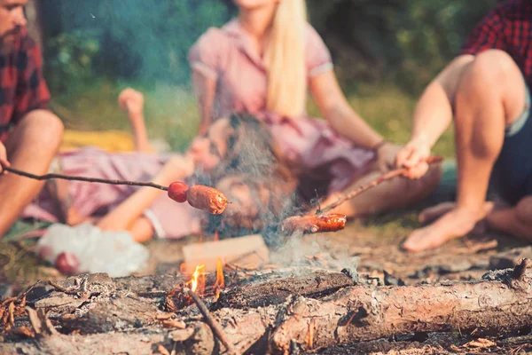 Cooking food on fire with blurred background of people. Sausages getting roasted on bonfire with smoke. Hot sausages on stick. Picnic or barbecue on nature. Camping food and meal concept