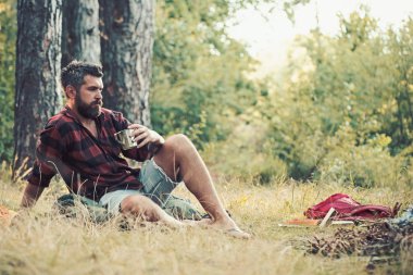 Hipster içecek çay veya kahve, şenlik ateşi. Sakallı adam suratına kamp ateşi ile rahatlayın. Adam ormanda kamp keyfini çıkarın. Yaz tatil kavramı. Kamp ve yürüyüş ve turistik