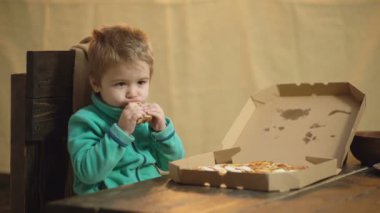 yeşil bir kazak çocuk ahşap bir sandalyede otururken bir pizza yiyor. Lezzetli pizza. Küçük çocuk pizza dilimi tutuyor. Aç çocuk pizzadan bir ısırık alıyor. Beslenme kavramı. Sağlıklı gıda konsepti.