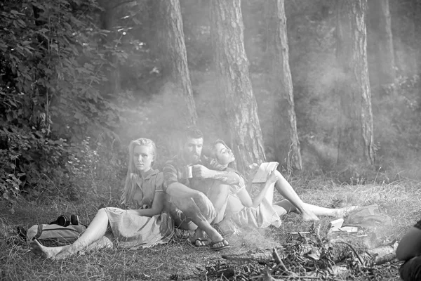 Comer comida, ler livros e entretenimento. Os amigos relaxam na fogueira com fumaça. Pessoas a acampar no fogo na floresta. Mulheres e homens barbudos na fogueira. Conceito de férias de verão, vintage — Fotografia de Stock