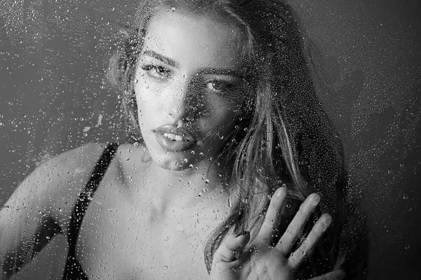 Gotas de lluvia en el cristal de la ventana con cara de niña. Ventana con gotas de agua antes de niña con maquillaje. Mujer sexy detrás de la hoja de plástico con gotas de agua. Moda y belleza. Ducha y tratamiento de spa higiene —  Fotos de Stock