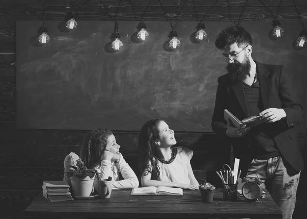 Δάσκαλος και τα κορίτσια τους μαθητές στην τάξη, chalkboard σε φόντο. Άνδρας με γενειάδα διδάσκει μαθήτριες, διαβάζοντας το βιβλίο. Περίεργος παιδιά χαρούμενα ακούγοντας το δάσκαλο με προσοχή. Δημοτικό σχολείο έννοια. — Φωτογραφία Αρχείου