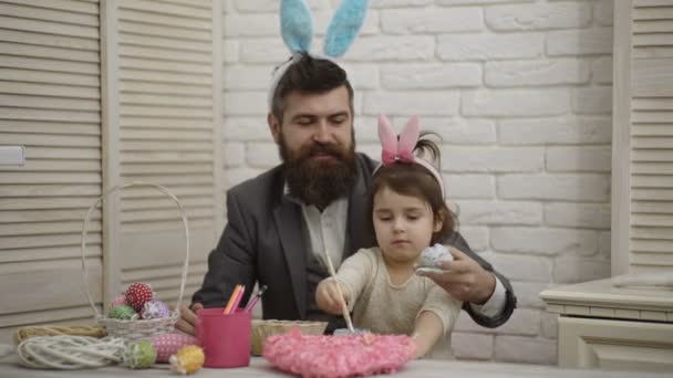 复活节快乐。爸爸和他的小女儿在为复活节假期做准备的时候玩得很开心。桌子上有复活节彩蛋、鲜花和油漆的篮子。美丽的女孩画复活节彩蛋. — 图库视频影像
