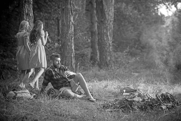 Hipster mit Bart sitzen am Lagerfeuer. bärtiger Mann trinkt Tee am Lagerfeuer mit Frauen auf verschwommenem Hintergrund. Freunde zelten gerne im Wald. Sommerferienkonzept. Zelten, Wandern und Reisen — Stockfoto