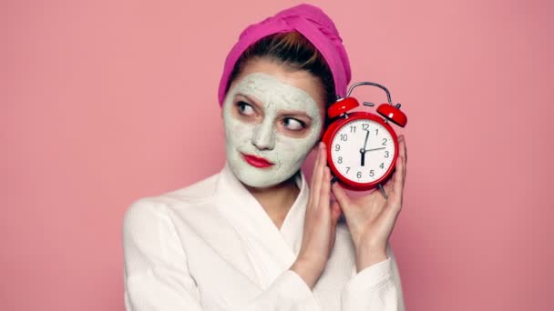女性はフェイスマスクを着ています。フェイスケアコンセプト美容トリートメント。Spa コンセプト。ウェルネスとスパのコンセプト。果物。ピンクの背景に彼女の手にマスクと目覚まし時計で驚いた女の子. — ストック動画
