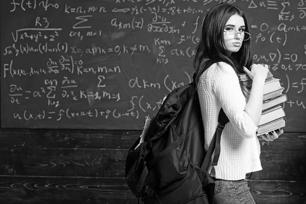 Brunette meisje permanent met zware stapel boeken en grote zwarte rugzak voor groen bord vol schrijven. Voorbereiding voor examen — Stockfoto