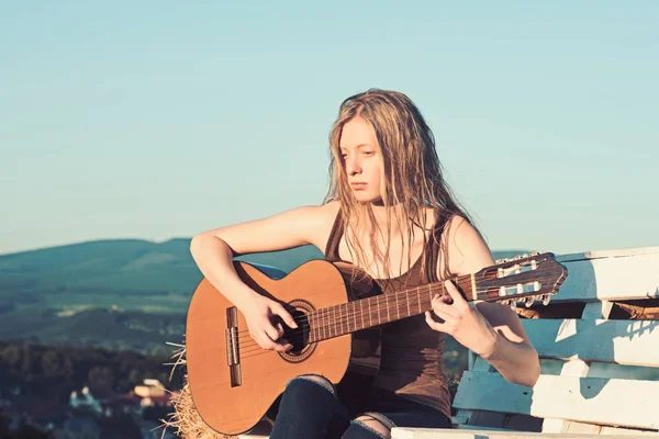 Albino girl hold acoustic guitar on blue sky. Albino woman play guitar on wooden bench on natural landscape