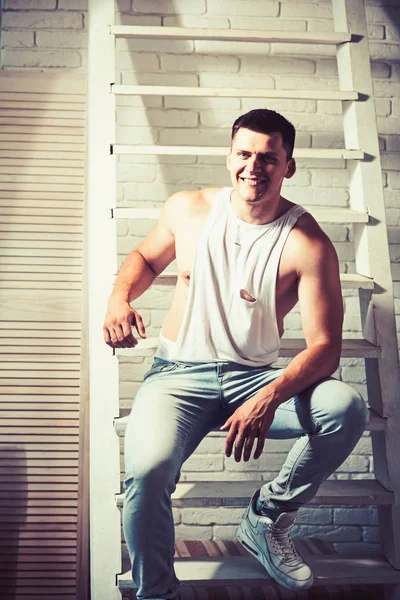 Un joven sonríe en la escalera. Chico en camiseta de moda y jeans de moda. Macho atlético con pecho y manos musculosas. Modelo de moda con cabello elegante. Estilo y tendencia de moda para hombre — Foto de Stock