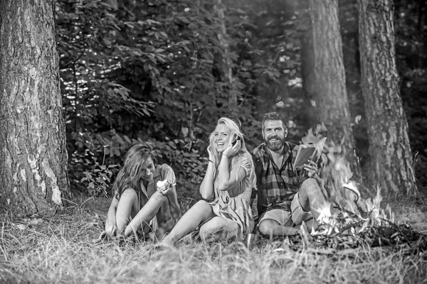 Amigos felizes se divertindo no piquenique na floresta. Homem barbudo com a namorada e a irmã sentadas perto da fogueira. Menina loira ouvindo música enquanto seu amigo está comendo maçã — Fotografia de Stock