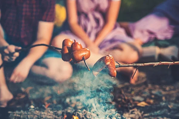 Ночевка в палатках летом. Готовить сосиски на палочках над пламенем костра. Две пары сидят у костра — стоковое фото