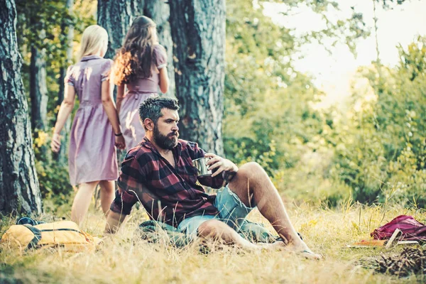 Un uomo barbuto che beve caffè o tè al falò. Due ragazze in abiti retrò che camminano sul sentiero nei boschi, tornano indietro. Concetti di relax e tranquillità — Foto Stock