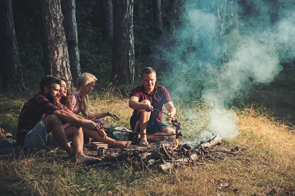 Dois casais a acampar na floresta. Cozinhar salsichas na fogueira. Noite quente de verão na floresta — Fotografia de Stock