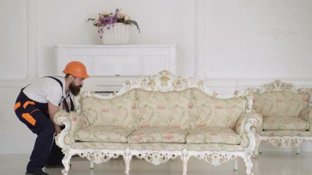 Τύπος παράδοσης σε προστατευτικό κράνος κινούμενο καναπέ σε ένα όμορφο λευκό εσωτερικό δωμάτιο. Ιδέα υπηρεσίας παράδοσης επίπλων. Νέοι μετακινούμενοι άντρας κινούμενο λευκό καναπέ σε νέο σπίτι με λευκό φόντο. — Αρχείο Βίντεο
