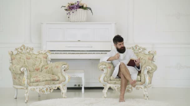 Homme en peignoir buvant du café et lisant un livre assis dans un fauteuil contre un piano blanc. Homme à la barbe et à la moustache profite du matin assis sur un fauteuil de luxe. Concept de loisirs Elite . — Video