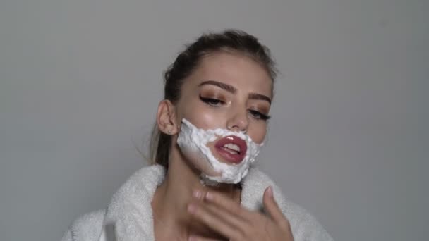 Woman with face covered with foam holds straight razor in hand. Girl on busy face wears bathrobe, grey background. Barber and shaving concept. Lady shaves her face with sharp blade of straight razor. — Stock Video