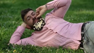 Yeşil çimençayırı üzerinde yatan papatya veya papatya çiçekleri ile çevrili mutlu gülümseyen adam. Neşeli yakışıklı sakallı adam güneşli bir gün açık havada zevk. Sakalında kamuflaj olan adam çimenlerin üzerinde yatıyor..