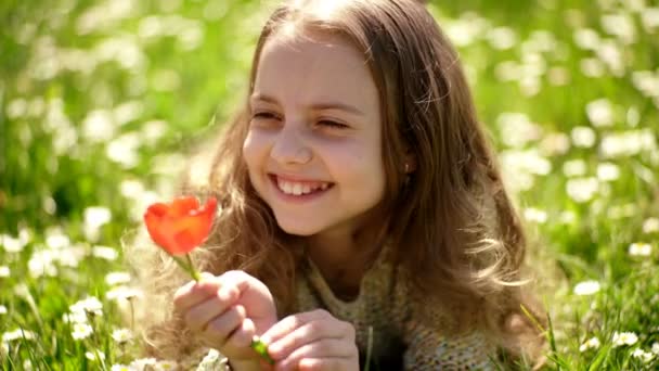 Meisje liggen op gras, grassplot op achtergrond. Tulip geur concept. Meisje op lachend gezicht heeft rode tulip bloem, geniet van de geur. Kind genieten van zonnige lente, liggend op de weide met bloemen. — Stockvideo