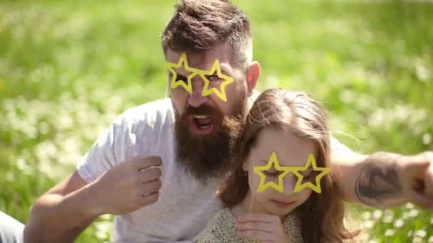 Concepto estrella de rock. Niño y padre posando con gafas en forma de estrella atributo fotomatón en el prado. Papá y su hija se sientan en la hierba en Grassplot, fondo verde. Familia pasar el ocio al aire libre . — Vídeos de Stock