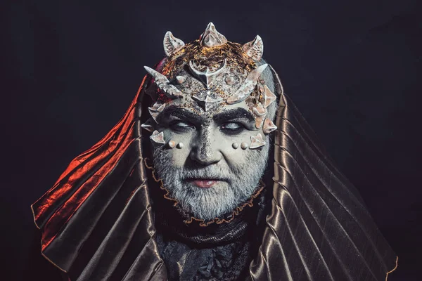 Senior man with white beard dressed like monster. Fantasy concept. Man with thorns or warts, face covered with glitters. Demon with golden hood on black background. Alien, demon, sorcerer makeup.