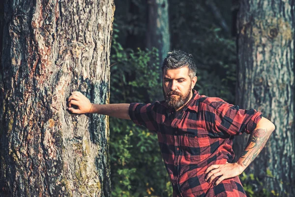 Großer brutaler Mann, der im Wald steht. Holzfäller, der sich an einen Baum lehnt. Hipster mit Rosen-Tattoo auf dem Arm — Stockfoto