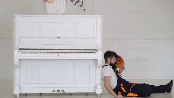 Loader lean on piano instrument. Man with beard, worker in overalls and helmet fall asleep tired, white background. Courier fall asleep while moving furniture, relocation. Tired worker concept. — Stock Video